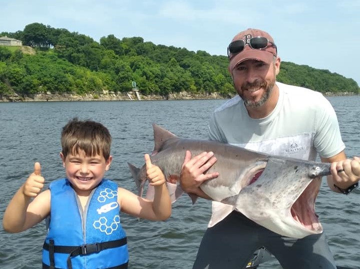 A young boy in a blue life jacket giving two thumbs up beside a smiling man holding a paddlefish, bluff shoreline behind them.