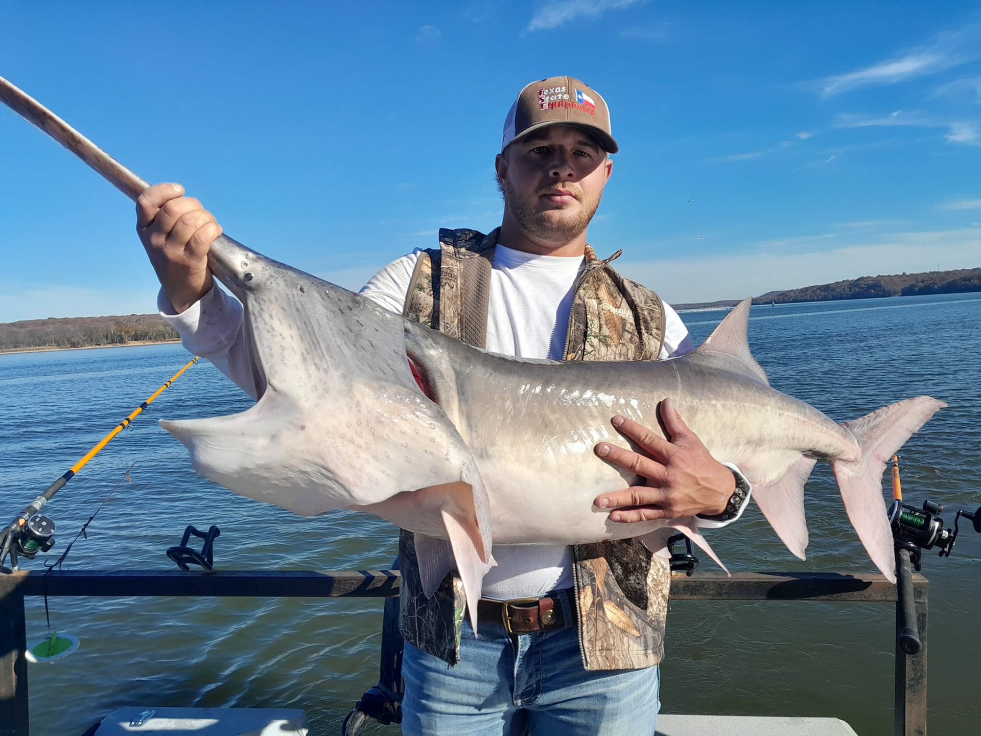 Angler standing in a boat holding a large paddlefish by the rostrum and body on an open Oklahoma lake under a clear blue sky.